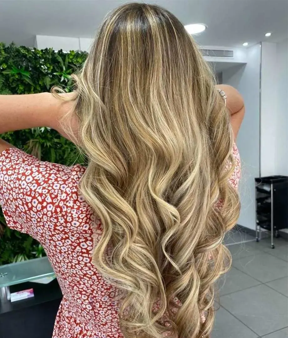 Balayage blond sur cheveux longs ondulés, devant le mur végétal du salon